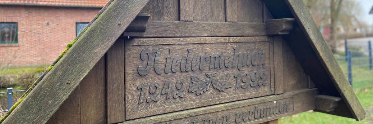 Ein hölzernes Schild auf dem der Name Niedermehnen eingeschnitzt ist. Im Hintergrund ist ein rotes Gebäude zu sehen. 