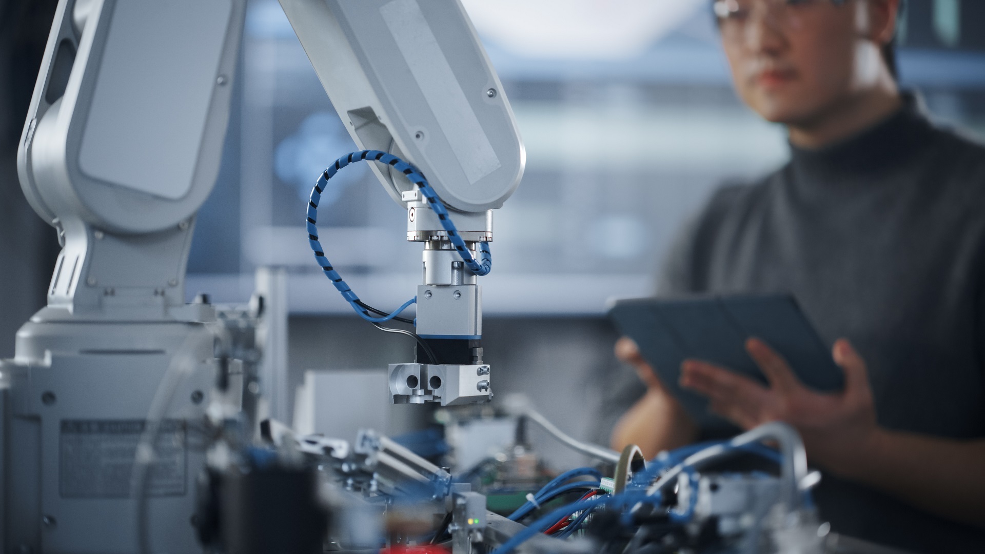 Ein Roboterarm in einem Industriebetrieb. Im Hintergrund ist eine Person zu sehen, die an einem Tablet Einstellungen vornimmt.