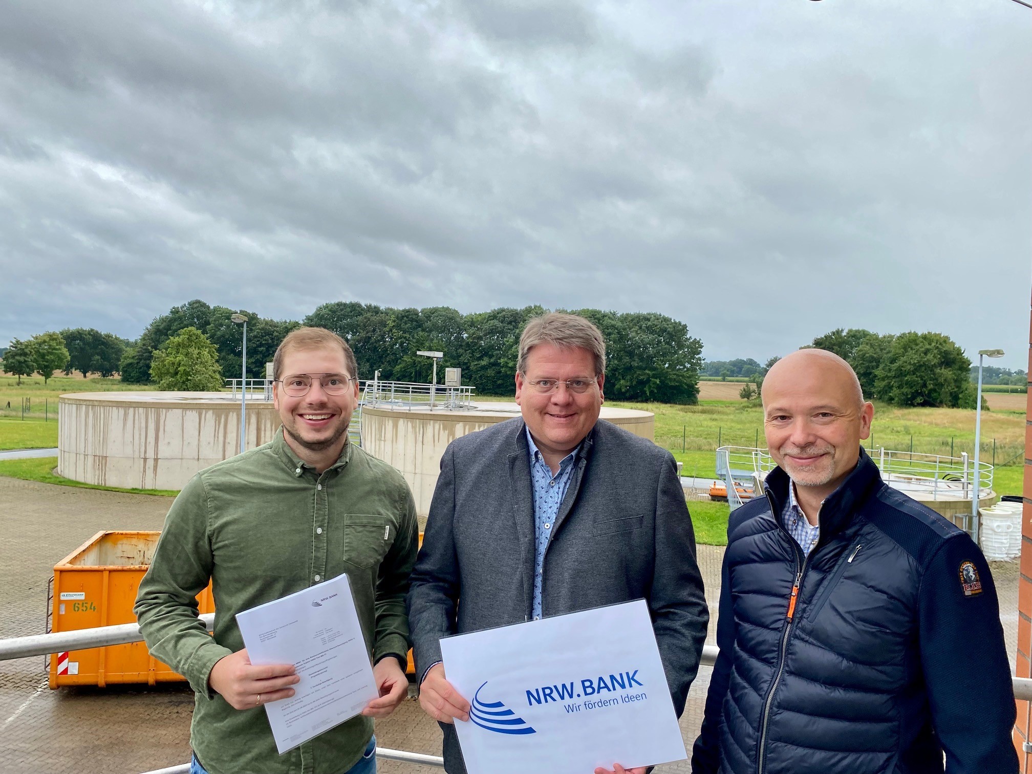 Drei Männer auf dem Gelände einer Kläranlage. Im Hintergrund sind eine Wiese sowie ein Klärbecken zu sehen. 