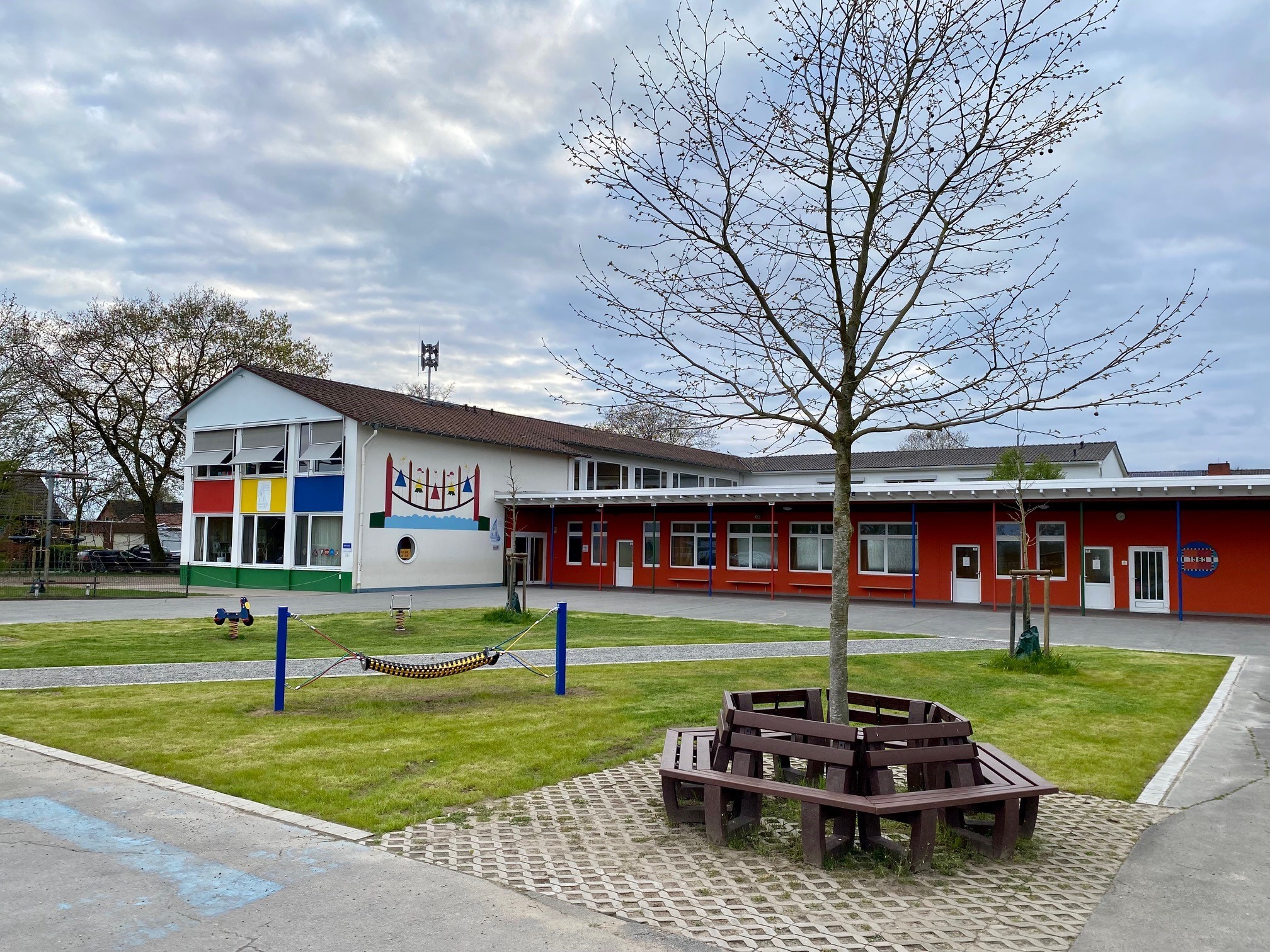 Ein begrünter Schulhof mit einer Rasenfläche, zwei Bäumen und einigen Spielgeräten, wie einem Schaukeltier und einer Hängematte. Im Hintergrund sind ein weißes und ein rotes Schulgebäude zu sehen. 