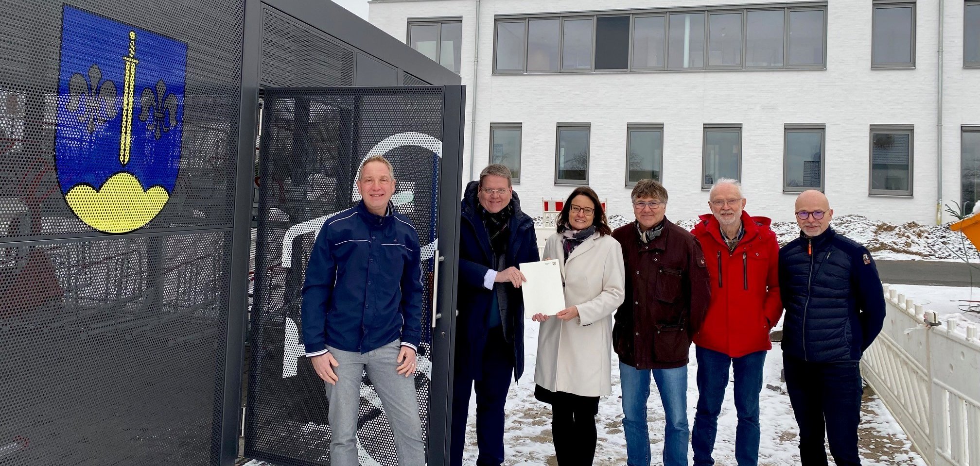 Gruppenfoto mit insgesamt sechs Personen, vor einer Fahrradgarage in einer verschneiten Landschaft. Eine Frau und ein Mann halten eine Urkunde in die Kamera.  