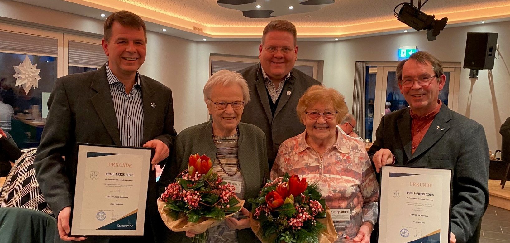 Die beiden Preisträgerinnen mit Blumenstrauß in der Hand, umrahmt von drei Männern, die alle in die Kamera lächeln.