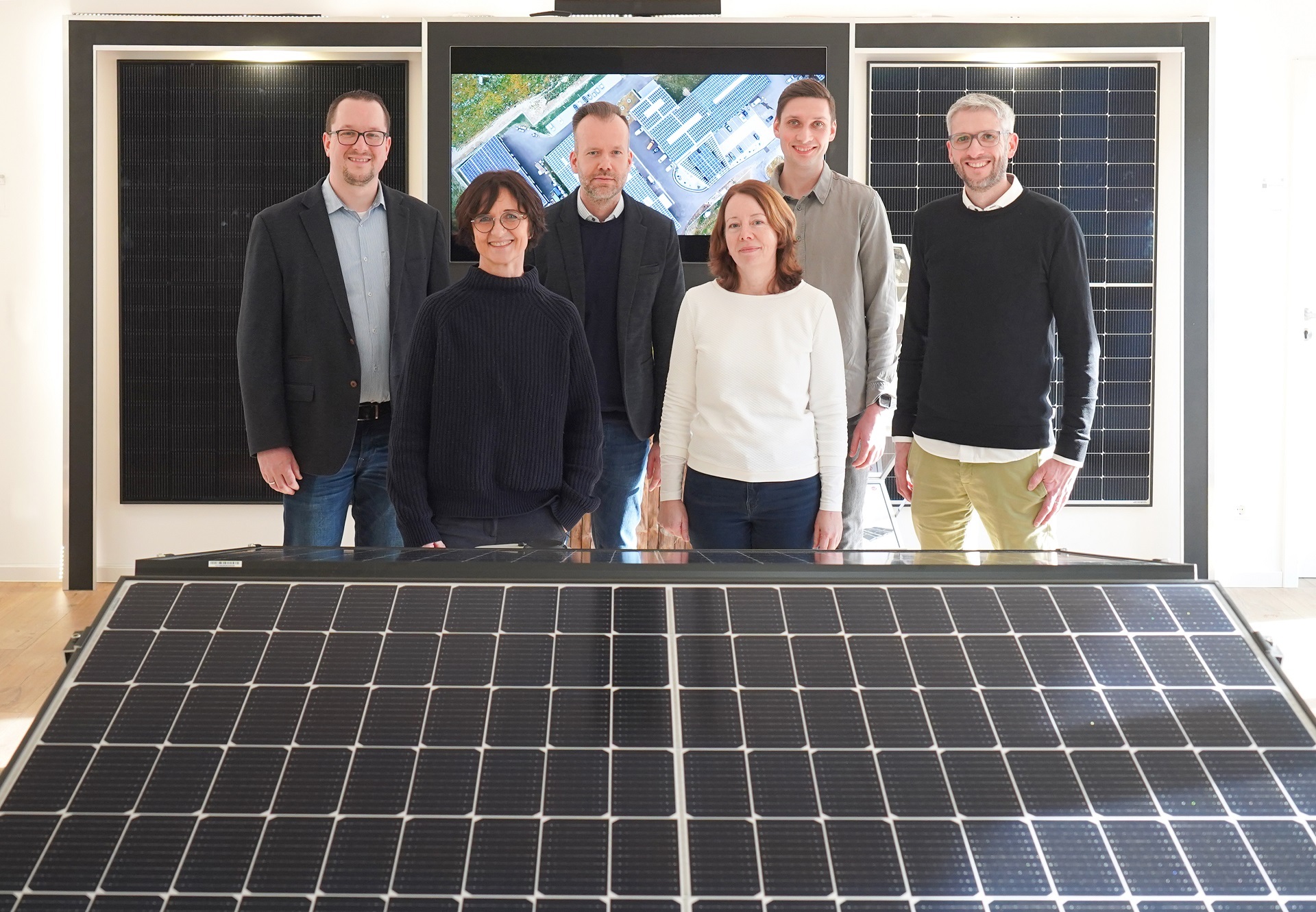 Zwei Frauen und vier Männer, die hinter einem Solarpanel stehen und in die Kamera lächeln.