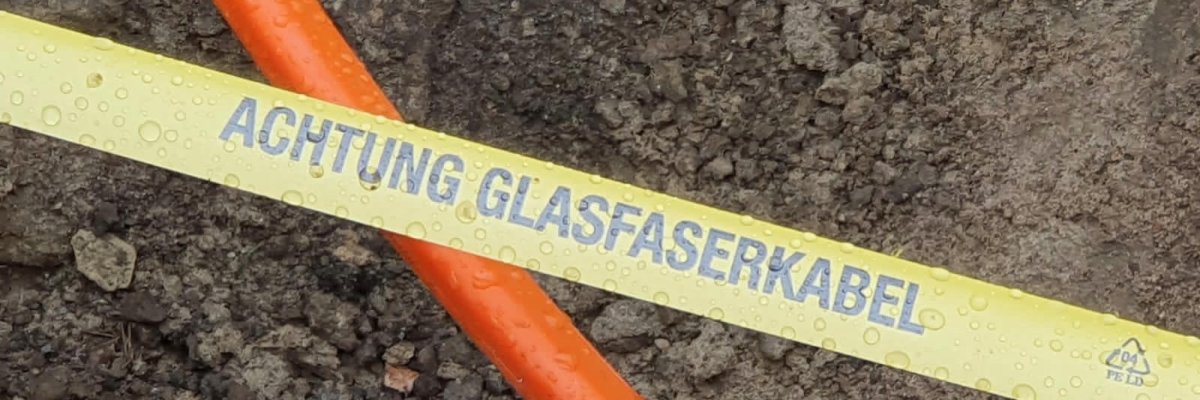 Achtung Glasfaser Ein oranges Kabel liegt in einer Baugrube. Eine Markierung zeigt, dass es sich um ein Glasfaserkabel handelt.