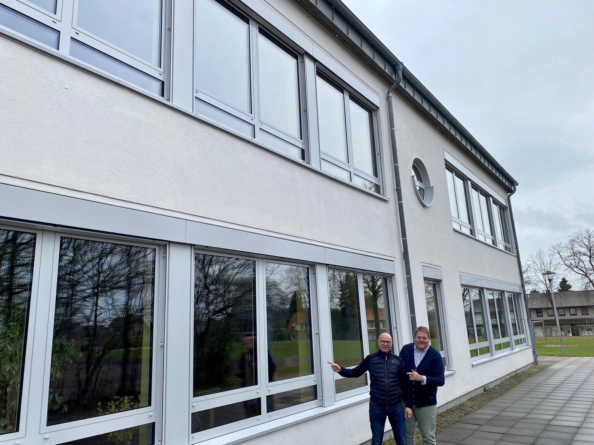 Zwei Männer stehen vor einer Fensterfront einer weißen Schulfassade. 