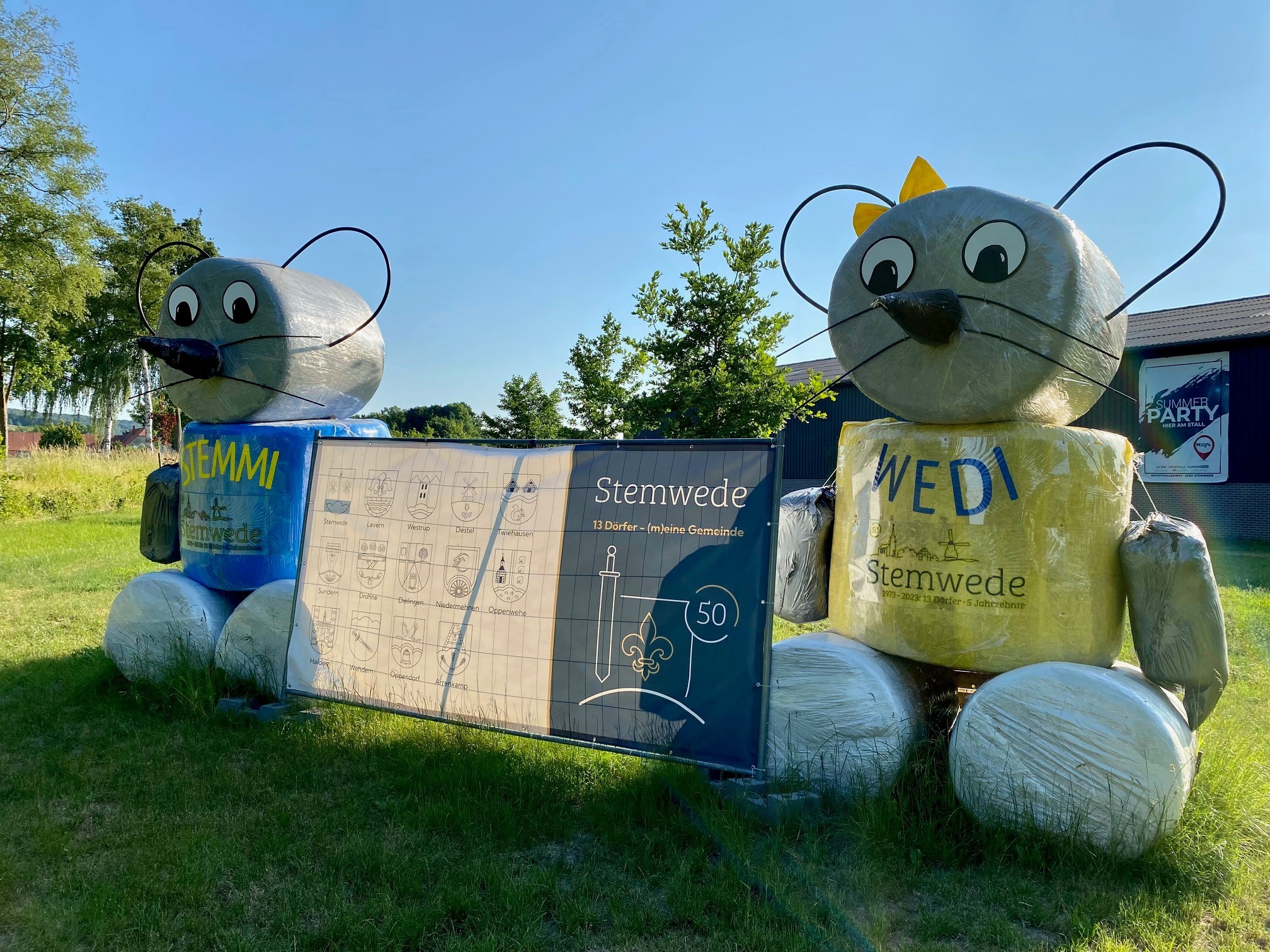 Auf einer Wiese stehen zwei Strohpuppen, die wie die beiden Maskottchenmäuse der Gemeinde Stemwede aussehen. In der Mitte der beiden Mäuse hängt ein blau-weißes Plakat, das auf das 50. Gemeindejubiläum der Gemeinde Stemwede hinweist. 