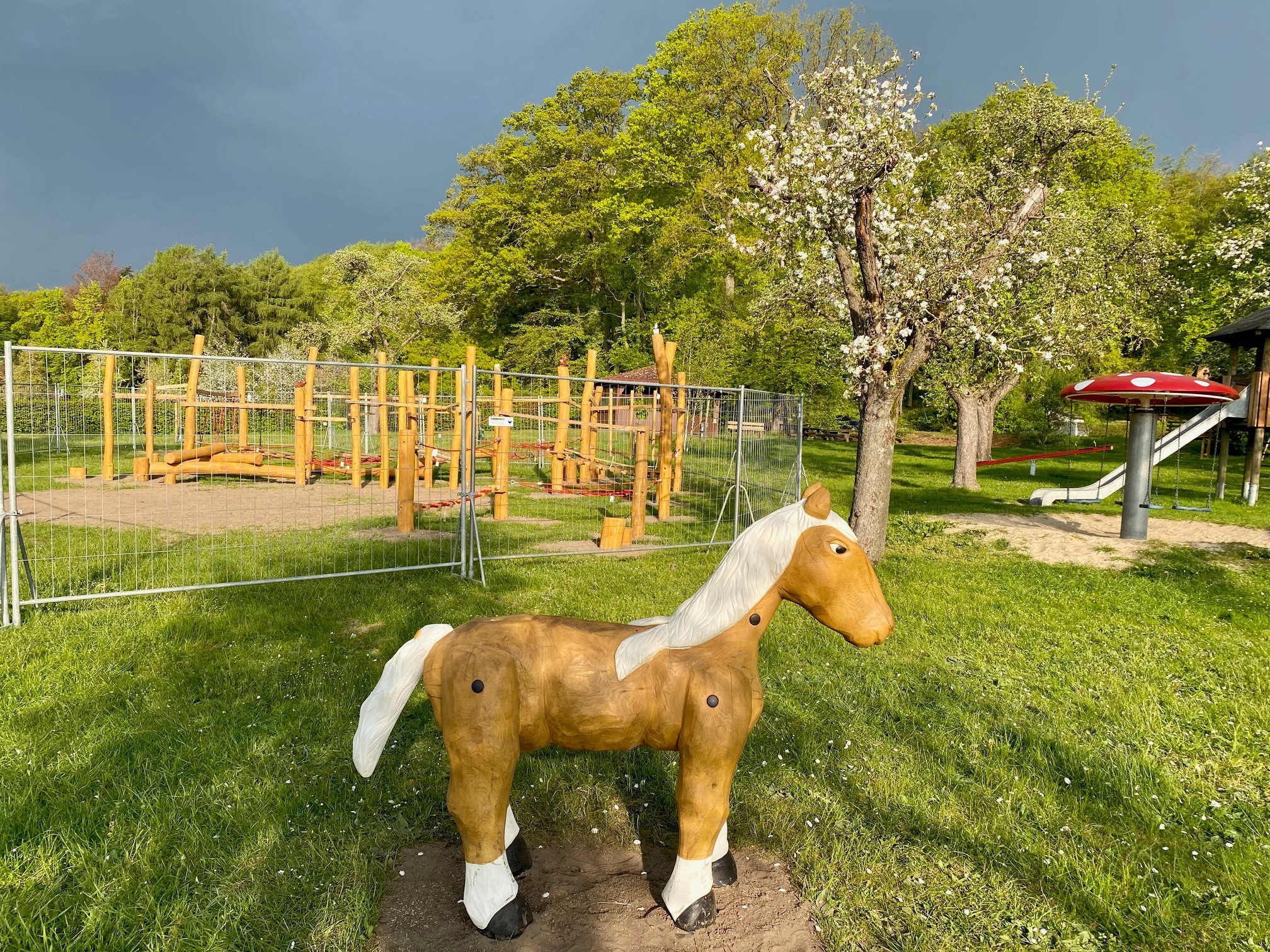 Ein Spielplatz auf dem im Vordergrund ein hölzernes Pferd zu erkennen ist und im Hintergrund ein eingezäuntes Spielmodul aus Holz. 