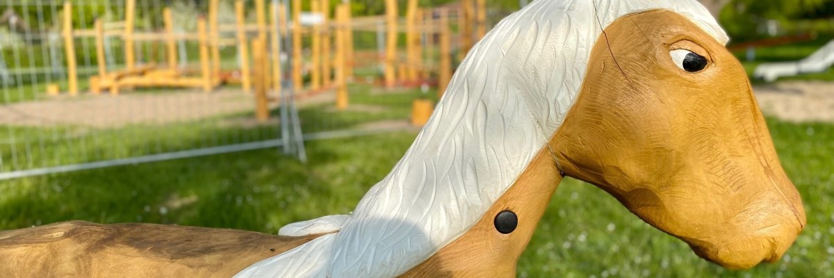 Großaufnahme eines hölzernen Pferdekopfes. Im Hintergrund ist die Silhouette eines Spielplatzes erkennbar.  