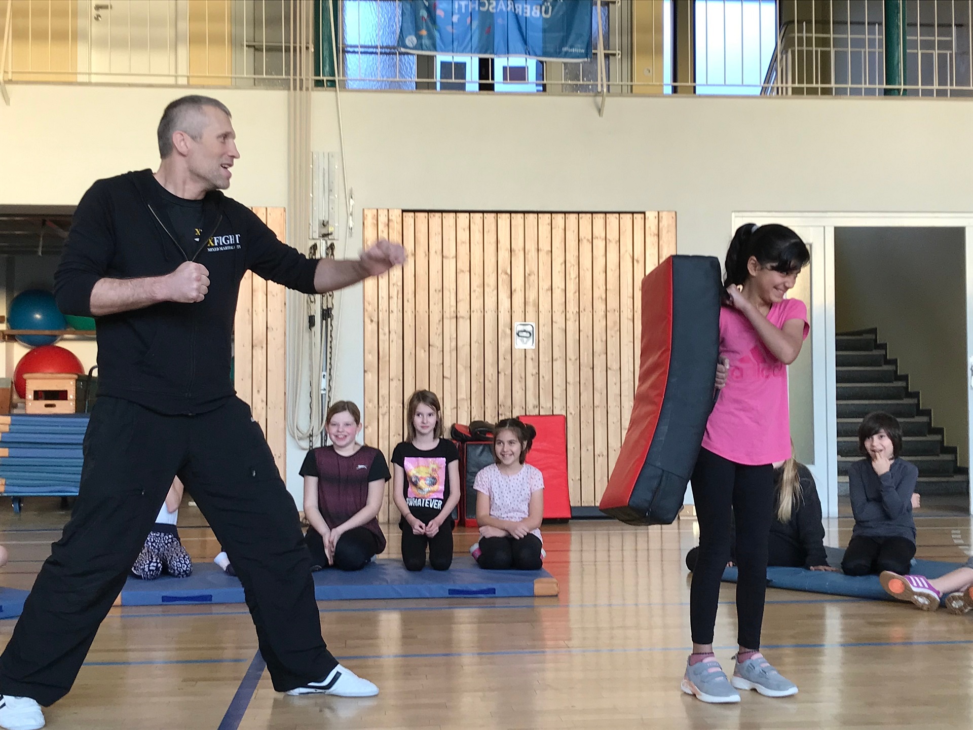 Ein kleines Mädchen hält ein dickes Kissen vor sich. Ein Mann in einem schwarzen Trainingsanzug steht vor ihr und ist kurz davor, gegen das Kissen zu treten. 