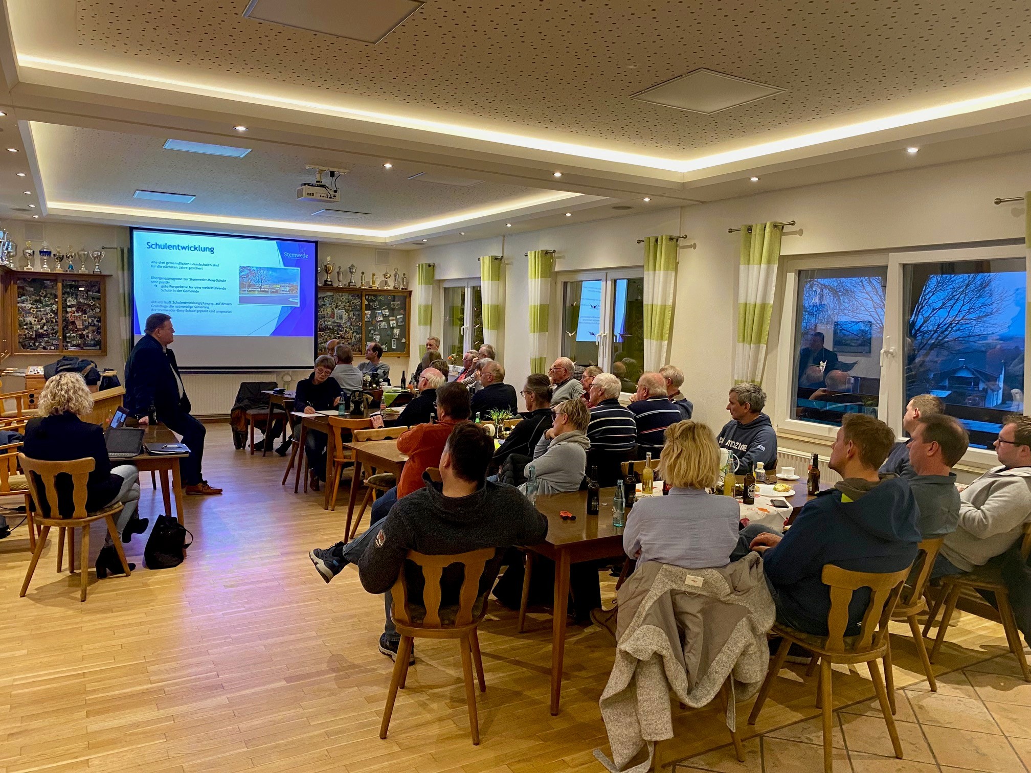 Ein Versammlungsraum, in dem etwa 30 Personen auf eine Leinwand schauen, auf der eine Präsentation gezeigt wird. 