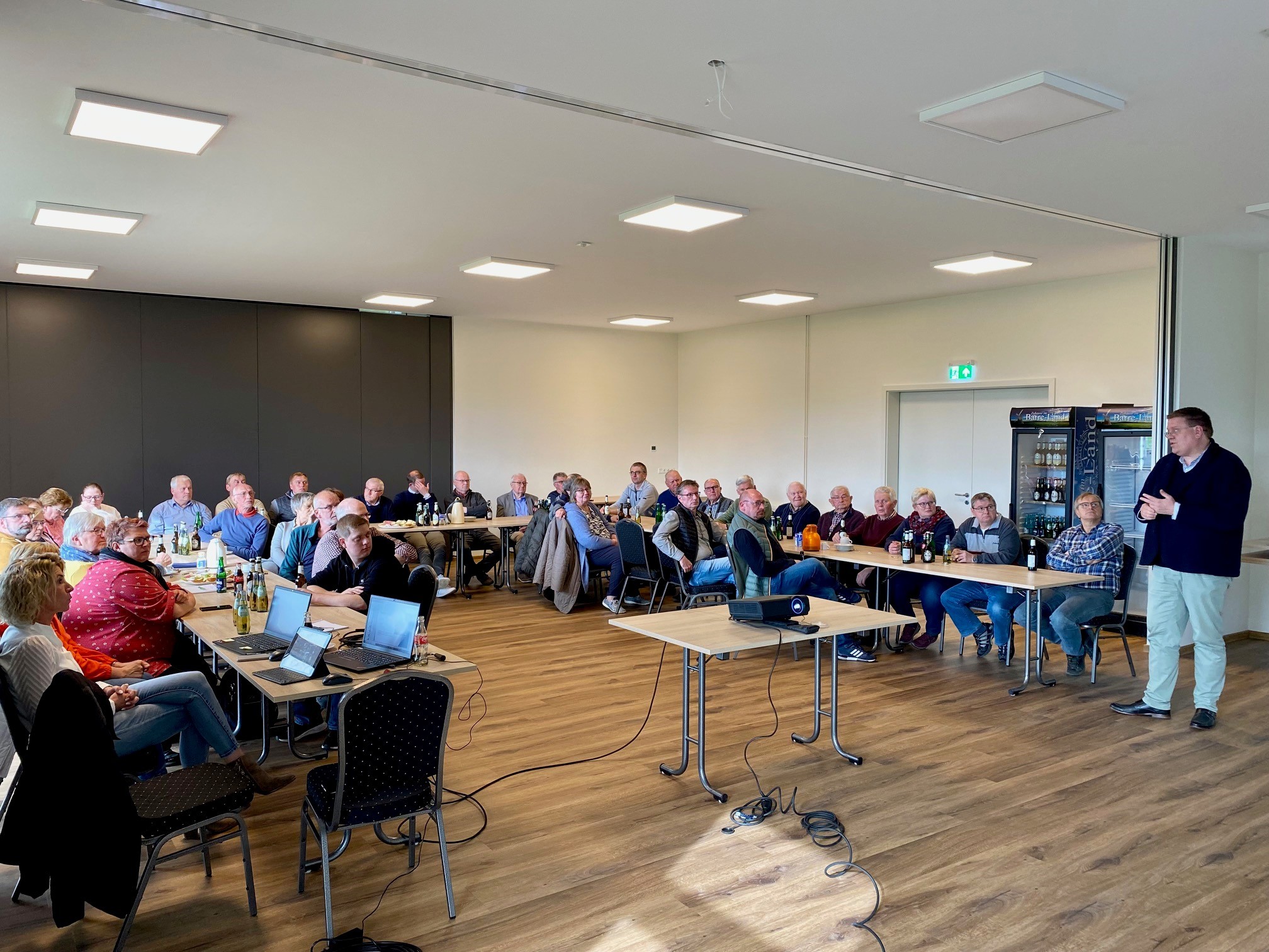 Ein Versammlungsraum in dem etwa 40 Leute an Tischen sitzen und einem Redner zuhören, der im rechten Bildrand steht. 