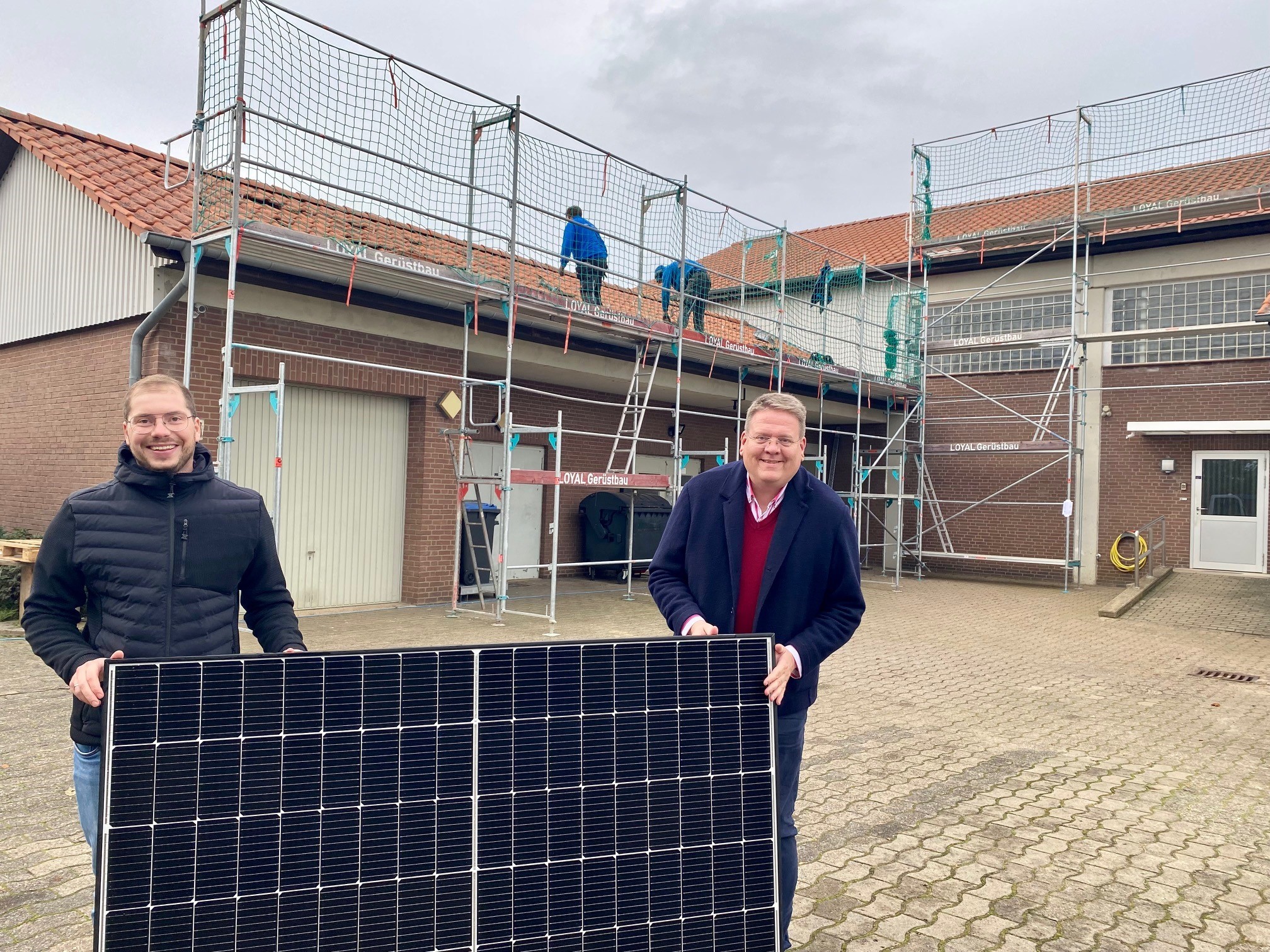 Zwei Männer, die ein Solarmodul festhalten. Im Hintergrund arbeiten Männer auf einem Gerüst, das an einem roten Gebäude steht. 