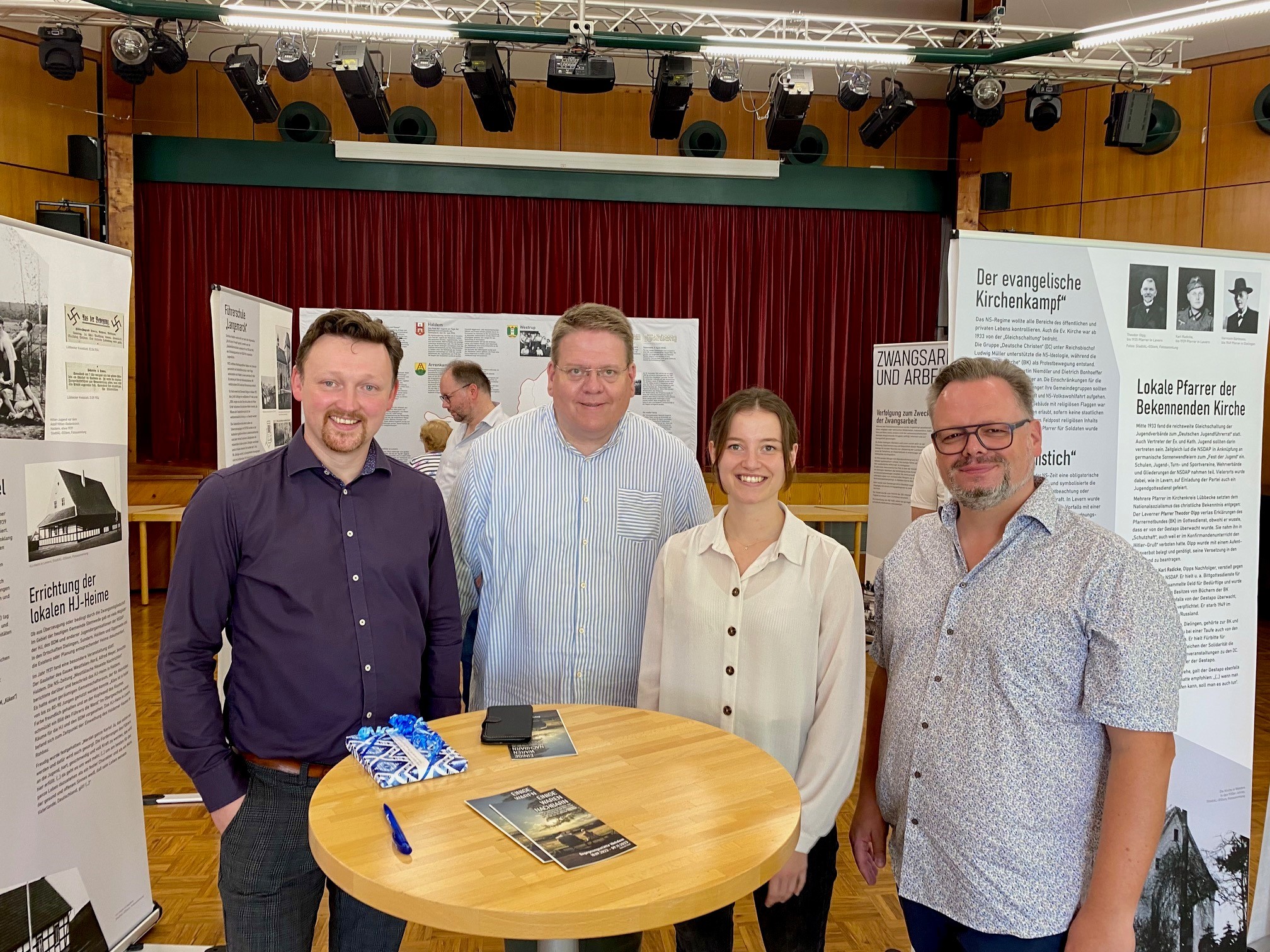 Vier Personen (eine Frau und drei Männer) in einem Ausstellungssaal. 
