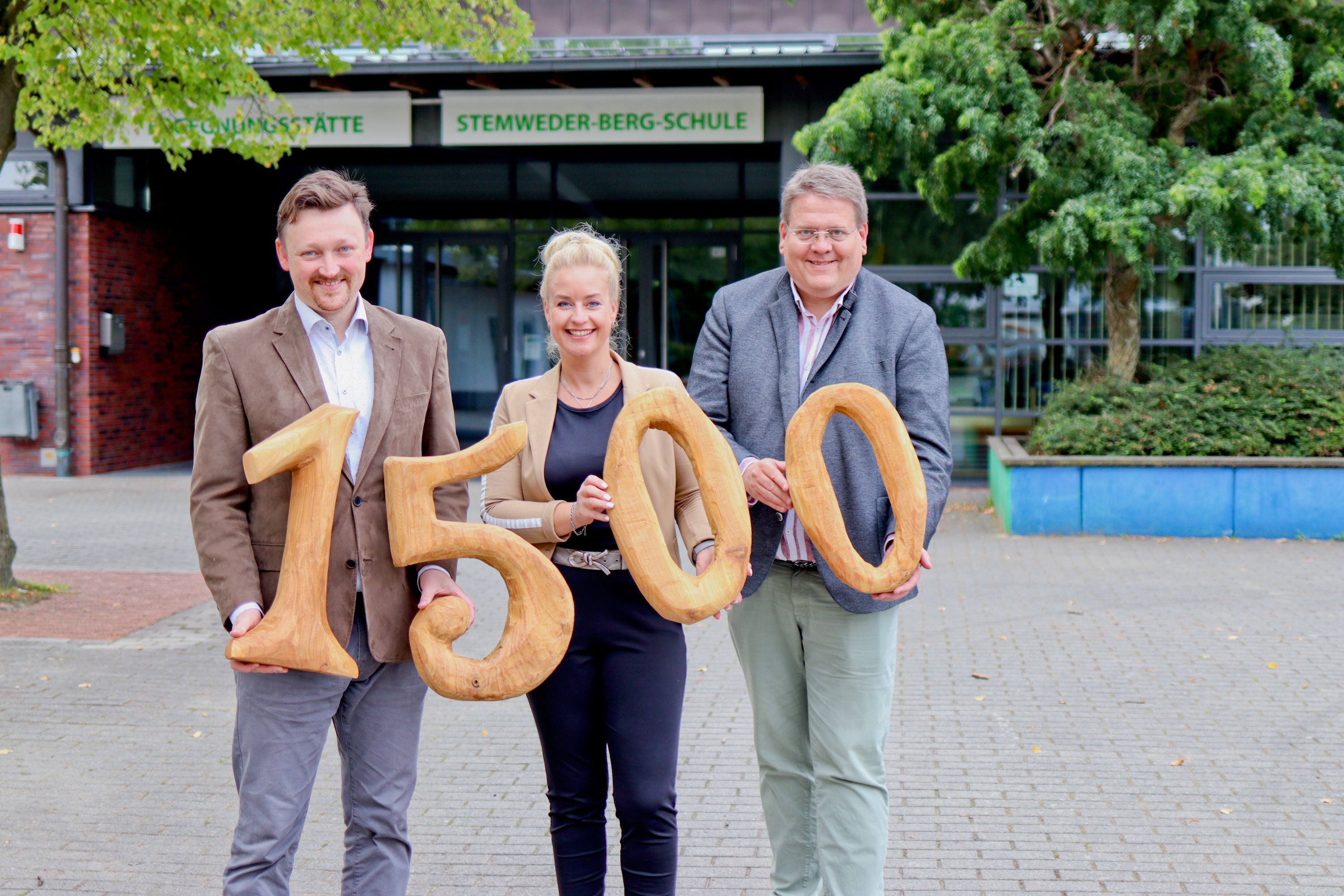 Drei Personen, zwei Männer links und rechts sowie eine Dame in der Mitte, stehen vor einem Schulgebäude und halten hölzerne Zahlen ins Bild, die die Spendensumme von 1.500 Euro symbolisieren. 