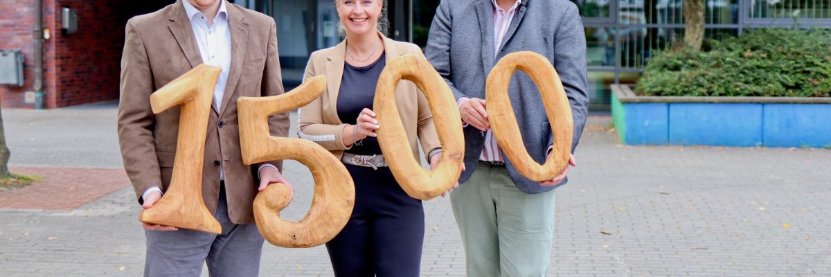 Drei Personen, zwei Männer links und rechts sowie eine Dame in der Mitte, stehen vor einem Schulgebäude und halten hölzerne Zahlen ins Bild, die die Spendensumme von 1.500 Euro symbolisieren. 