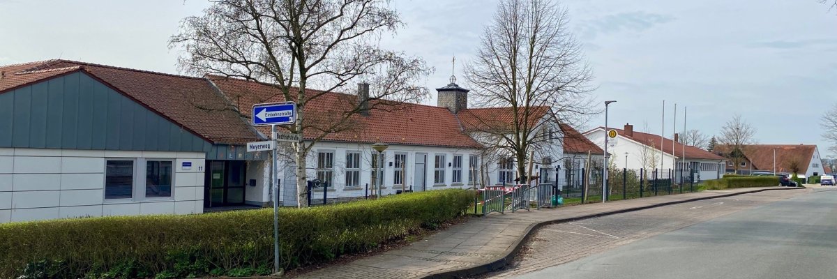 Ein weißes Schulgebäude mit einer davor verlaufenden Bushaltestelle an einer Straße. 