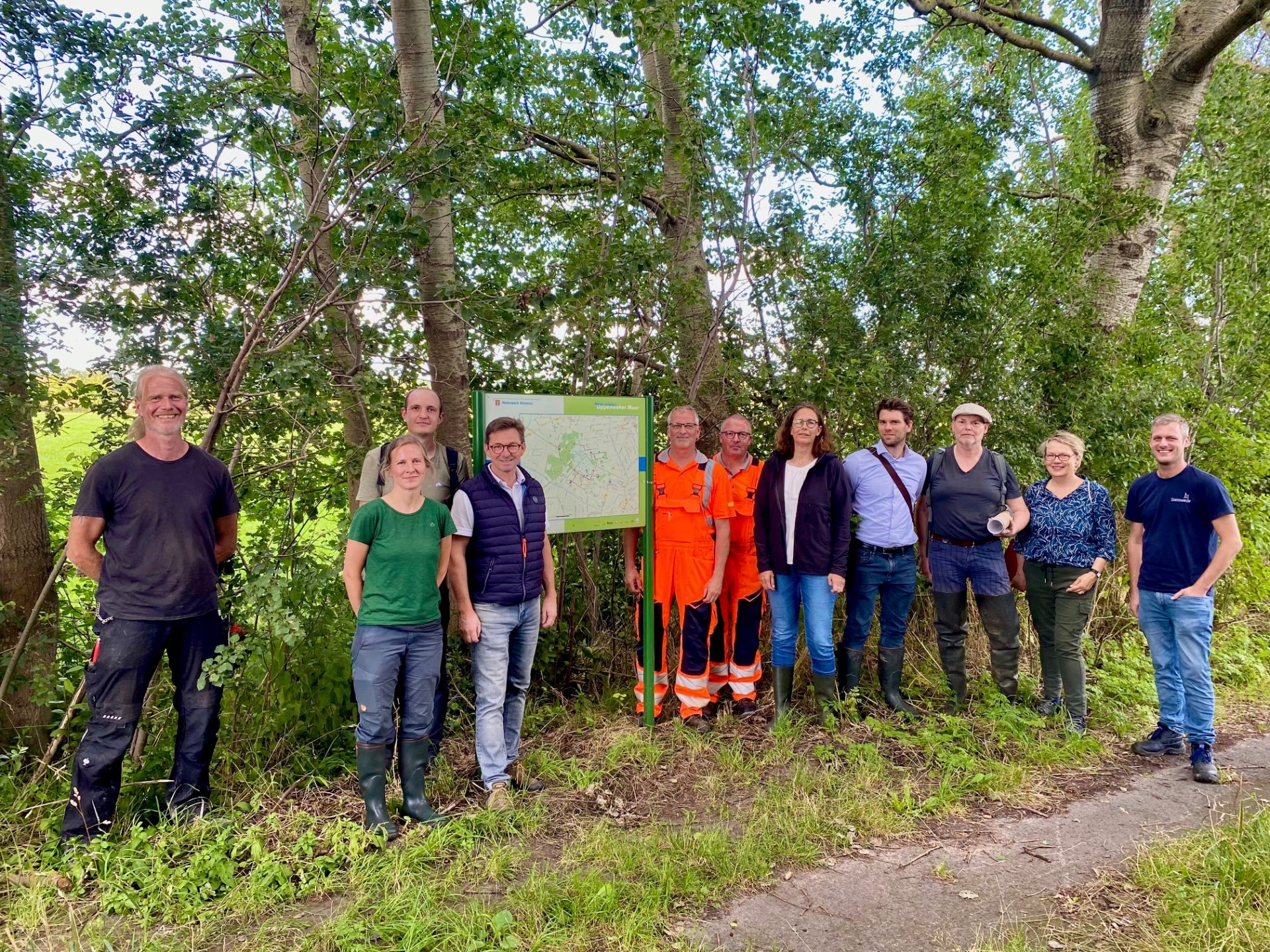 Mehrere Personen stehen vor einer Wanderkarte, die auf einem Grünstreifen steht.  