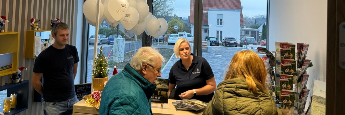 Ein Verkaufsraum, in dem eine Frau in einem dunkelblauen Polo-Shirt zwei Kunden bedient. Im Hintergrund sind Ballons und einiges an Deko zu sehen.