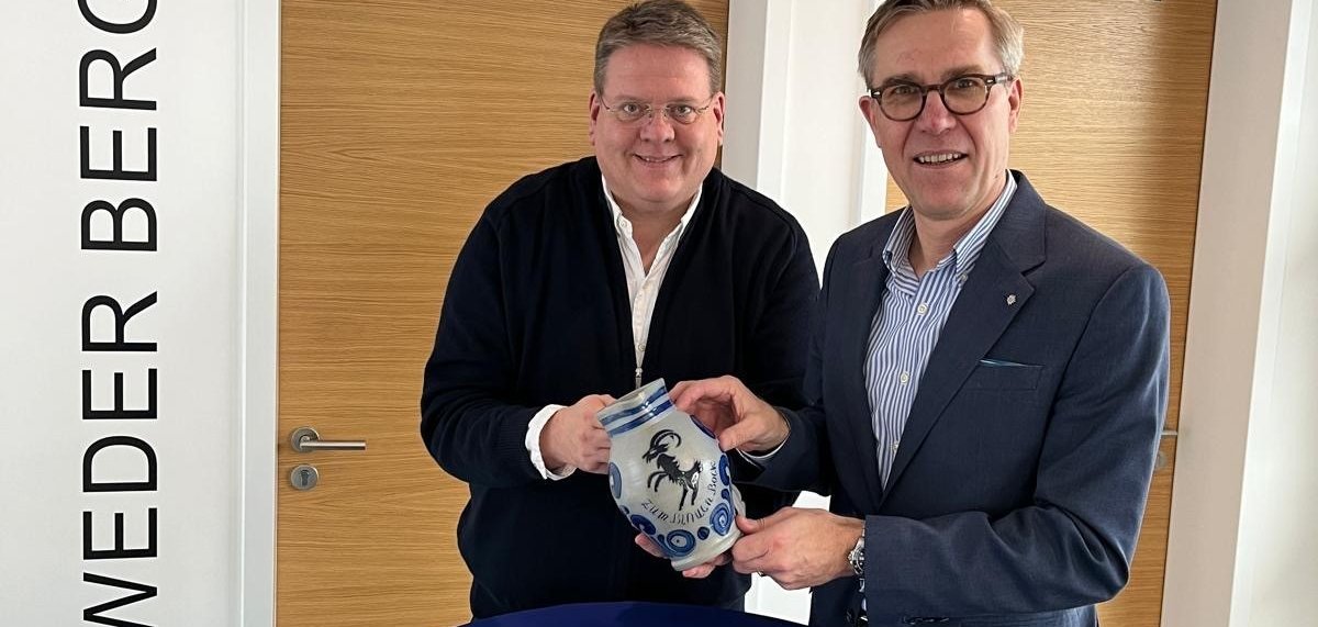 Zwei Männer stehen an einem blauen Stehtisch und halten einen grauen Steinkrug in die Kamera, der blaue Verzierungen, einen blauen Springbock und die Aufschrift "Zum Blauen Bock" zu sehen sind.