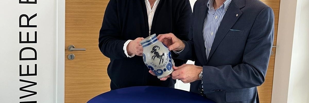 Zwei Männer stehen an einem blauen Stehtisch und halten einen grauen Steinkrug in die Kamera, der blaue Verzierungen, einen blauen Springbock und die Aufschrift "Zum Blauen Bock" zu sehen sind.