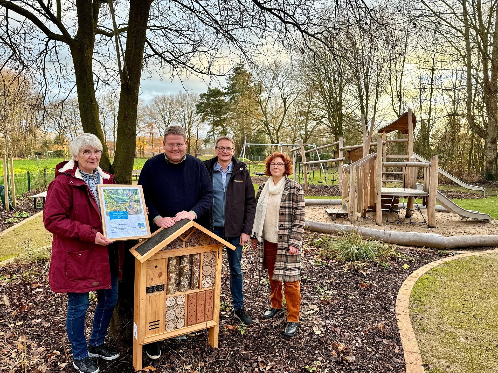 Zwei Männer und zwei Frauen stehen in einer herbstlichen Parkanlage. Ein Mann hält ein Insektenhotel, die Frau links im Bild eine eingerahmte Urkunde. Im Hintergrund ist ein Spielgerüst aus Holz zu erkennen.