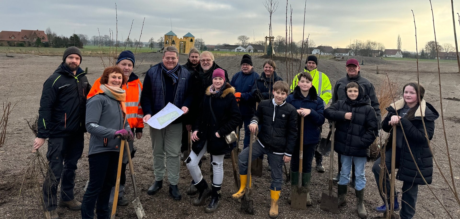 Eine Gruppe von Erwachsenen und Jugendlichen in wetterfester Kleidung und zum Teil mit einem Spaten in der Hand, auf einem weitläufigen Areal aus Mutterboden. Hier entsteht ein Spielplatz.