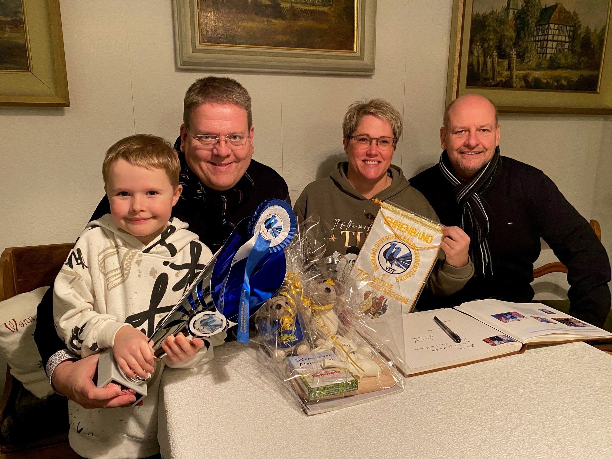 Ein Kind hält einen Siegerpokal in den Händen. Rechts neben ihm sitzen zwei erwachsene Männer und eine Frau. 