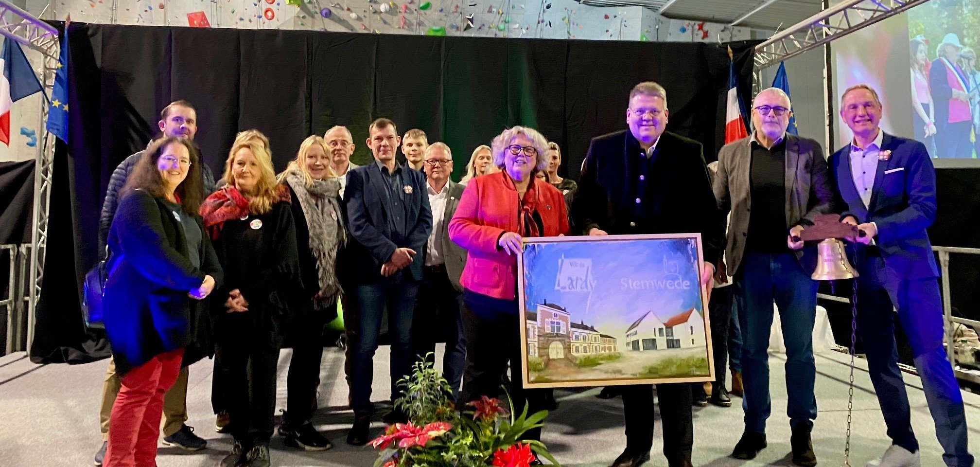 Ein Gruppenfoto mit mehreren Erwachsenen  auf einer Bühne. Eine Frau und ein Mann halten ein selbst gemaltes Bild auf dem Häuser und ein Schriftzug zu sehen sind.