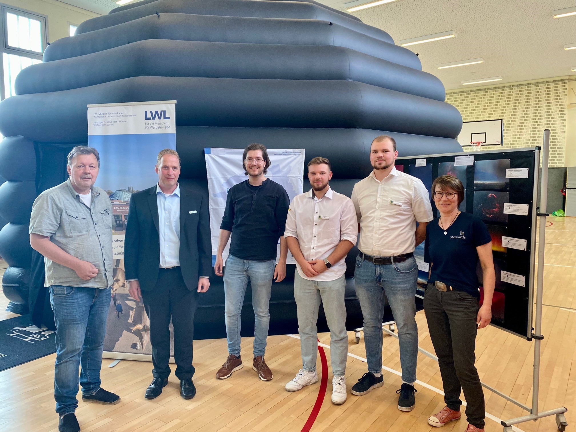 Fünf Herren und eine Frau stehen in einer Turnhalle und lachen in die Kamera. Im Hintergrund ist eine große, schwarze aufblasbare Kuppel zu erkennen sowie eine Stellwand, an der Fotos von Sternenaufnahmen hängen. 