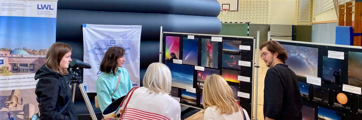 Planetarium 2024-03 Eine Gruppe von Frauen lässt sich von einem braunhaarigen Mann mit Bart Fotoaufnahmen erklären, die an einer Stellwand hängen und Aufnahmen vom nächtlichen Himmel zeigen. Die Stellwand steht in einer Turnhalle. Im Hintergrund ist eine schwarze, aufblasbare Kuppel zu sehen.