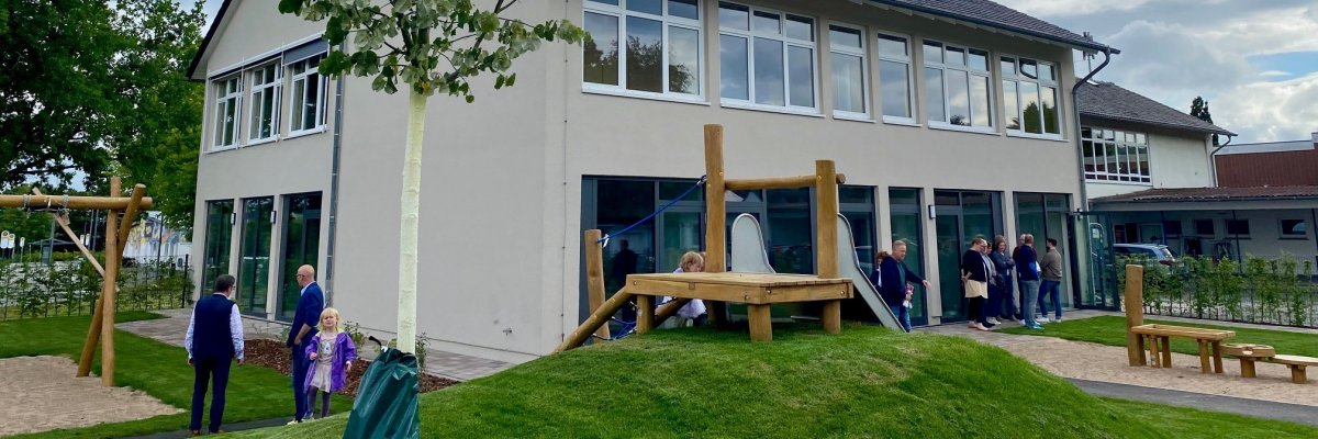 Eine Aufnahme eines beigen, zweistöckigen Gebäudes mit vielen Fenstern. Vor dem Gebäude ist ein Spielplatz zu erkennen - das Außengelände eines Kindergartens.