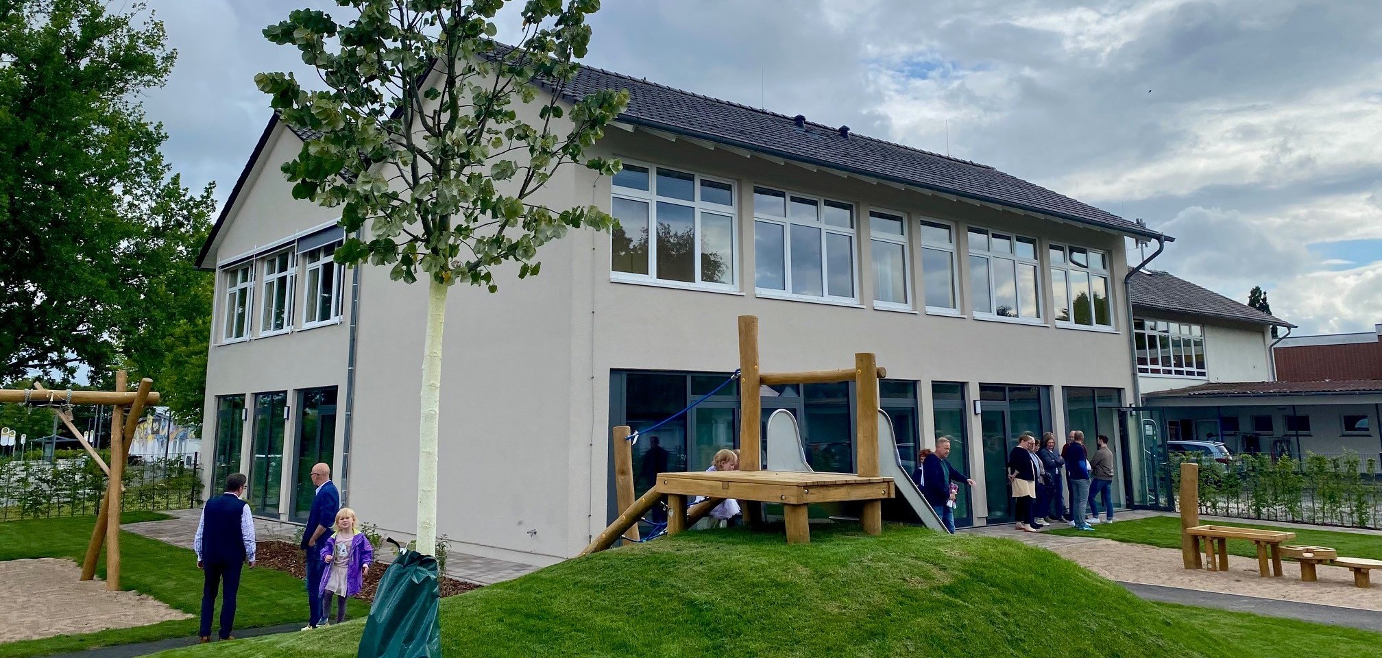 Eine Aufnahme eines beigen, zweistöckigen Gebäudes mit vielen Fenstern. Vor dem Gebäude ist ein Spielplatz zu erkennen - das Außengelände eines Kindergartens. 