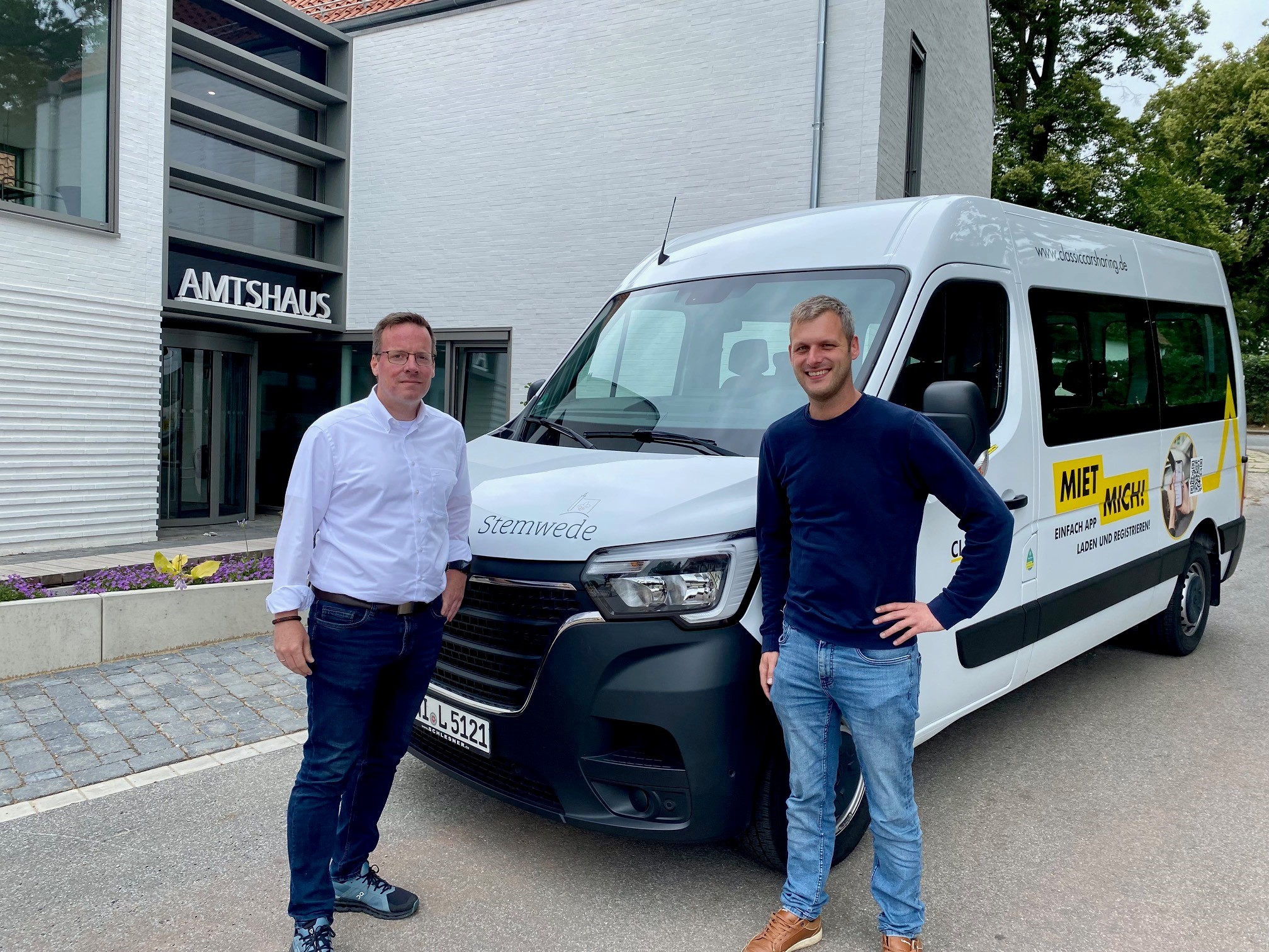 Vor der Fassade eines modernen, weißen Gebäudes, an dem die Aufschrift "Amtshaus" zu lesen ist, steht ein weißer Bulli mit der Aufschrift "CarSharing Stemwede". Vor dem Bulli stehen zwei Männer, die in die Kamera lächeln.  