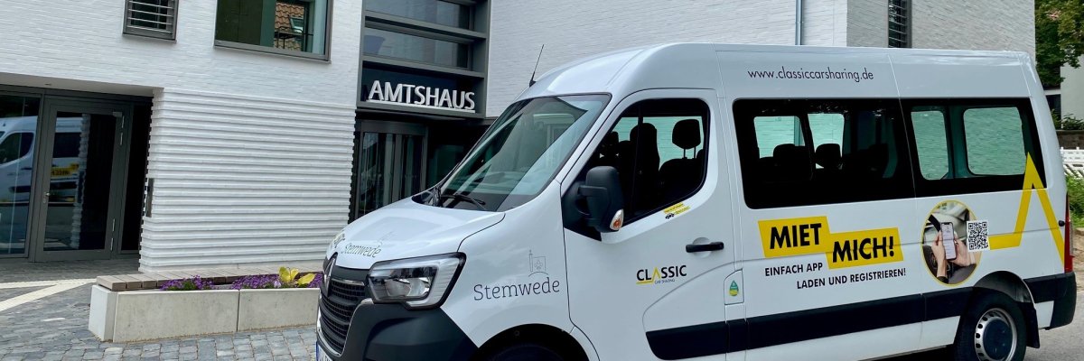 Vor der Fassade eines modernen, weißen Gebäudes, an dem die Aufschrift "Amtshaus" zu lesen ist, steht ein weißer Bulli mit der Aufschrift "CarSharing Stemwede".