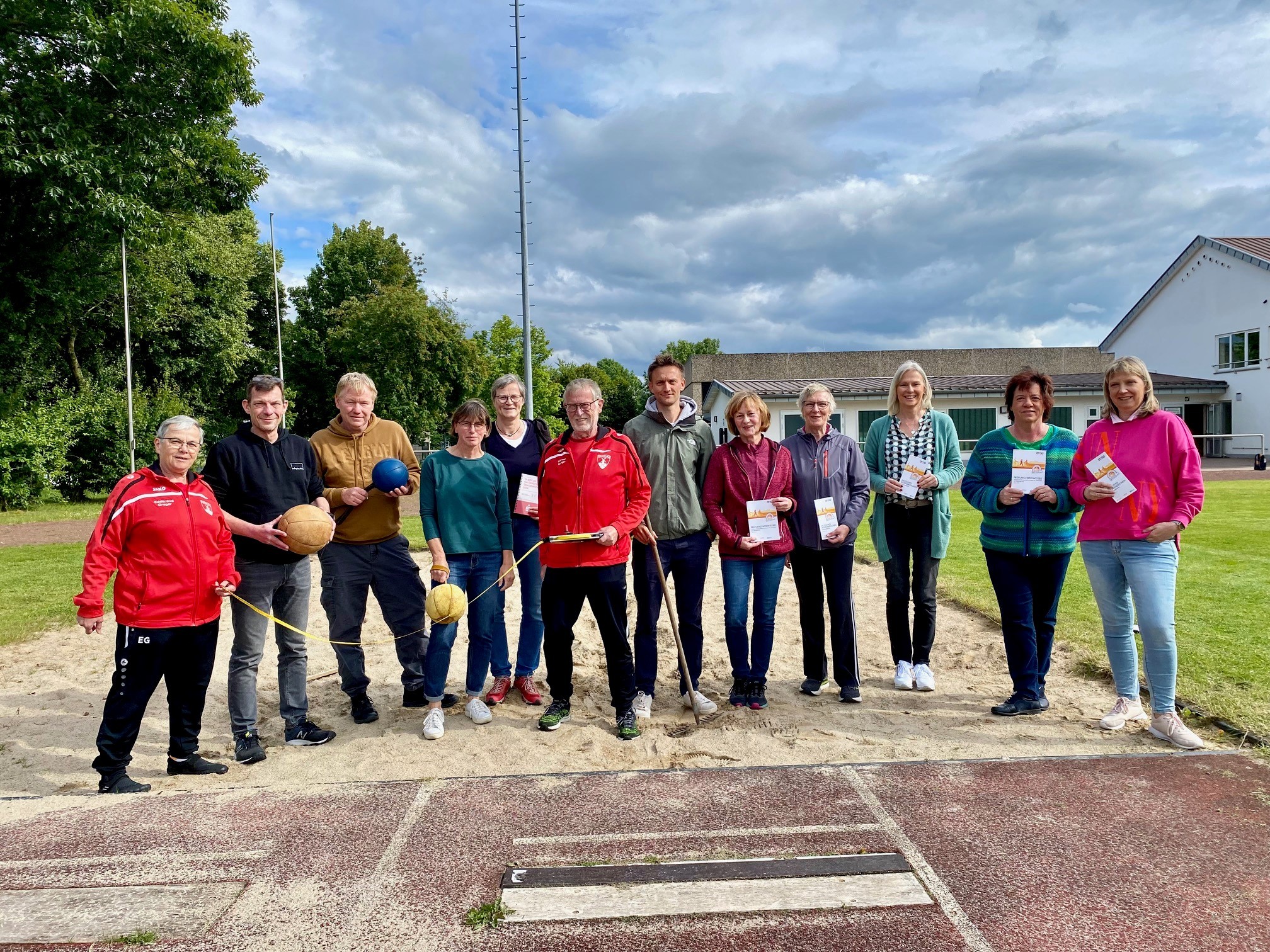 Insgesamt 12 Personen, 8 Frauen und 4 Männer, stehen auf einem Sportplatz im Sand einer Sprungkuhle und halten verschiedene Sportgeräte, die für das Sportabzeichen benötigt werden, in den Händen. Zum Beispiel Schlagbälle, Metermaß oder auch eine Harke.