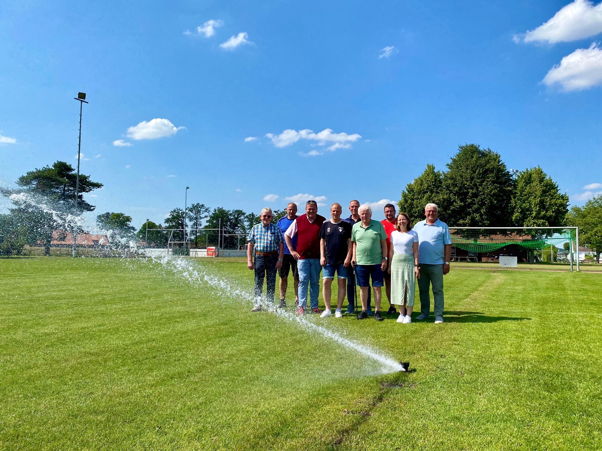 Eine Gruppe von Menschen steht an einem sonnigen Tag auf einem Sportplatz. Vor ihnen schaut eine Rasensprengerdüse aus dem Boden, die in Betrieb ist und Wasser ausstößt.