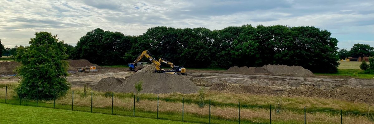 Blick auf eine große, ausgebaggerte Fläche einer Baustelle. Ein gelber Bagger sowie ein Trecker mit Anhänger sind zu sehen.