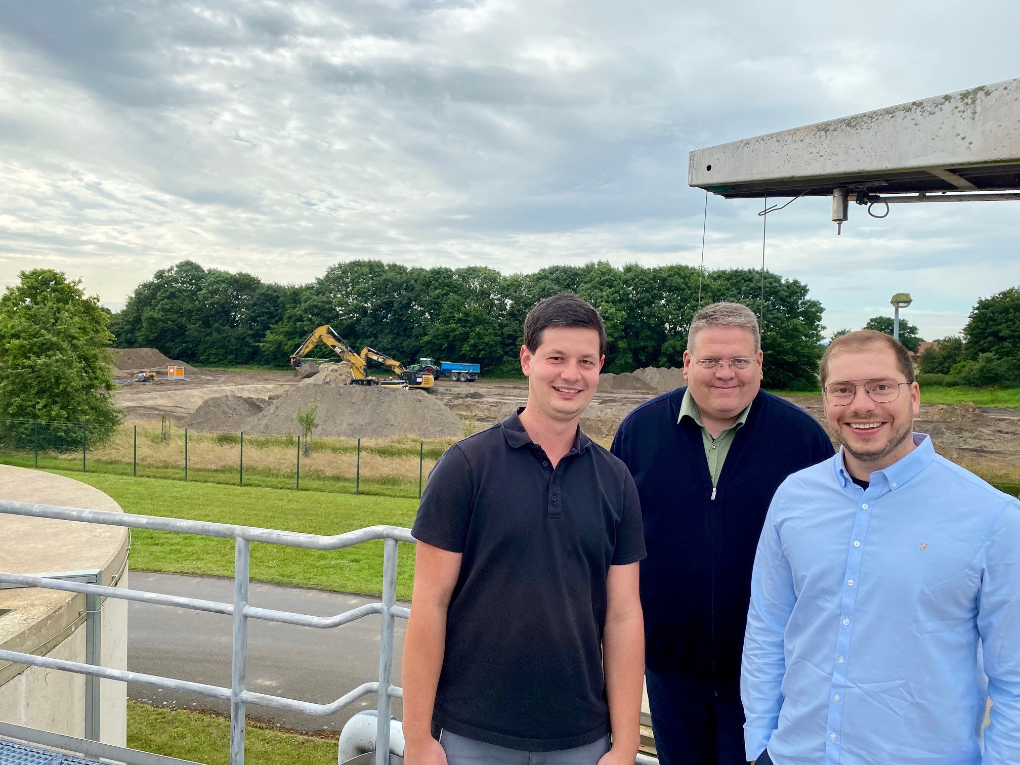 Drei Männer, die vor einer ausgebaggerten Fläche einer großen Baustelle stehen und in die Kamera lächeln.