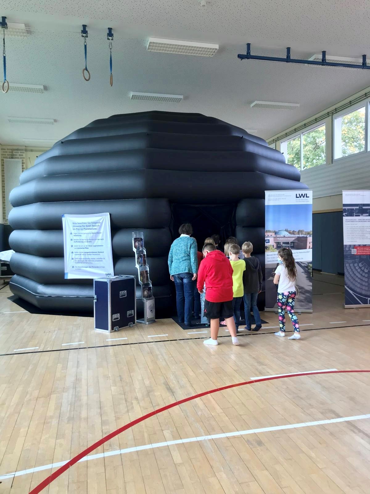 Eine schwarze, runde aufblasbares Kuppel, steht in einer Turnhalle. Eine Dame und mehrere Kinder schauen gerade in das Innere der Kuppel. 
