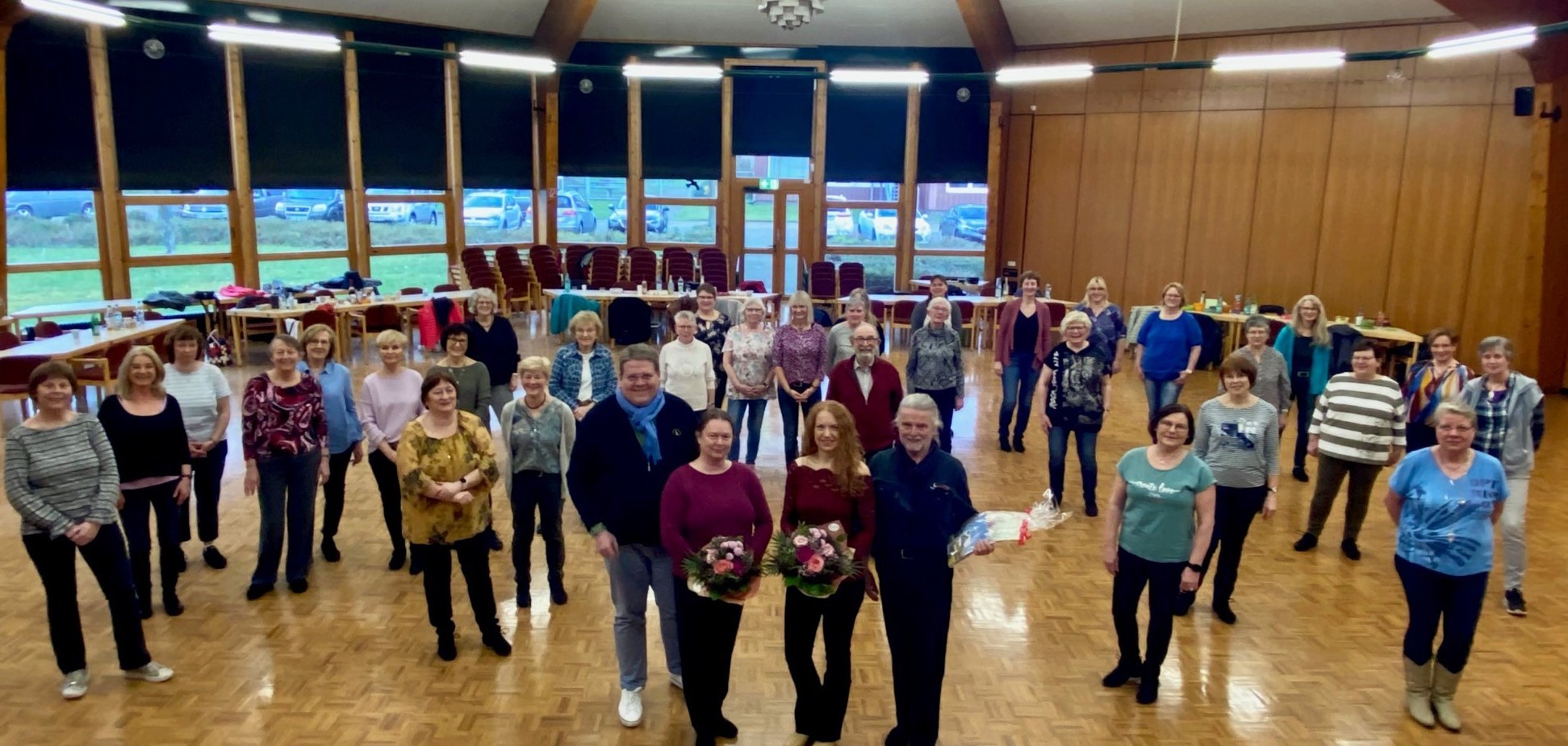 Zahlreiche Erwachsene, die verteilt in einem Veranstaltungsaal stehen und in die Kamera schauen. Im Vordergrund stehen zwei Frauen und zwei Männer, die Blumen oder Präsente in den Händen halten.