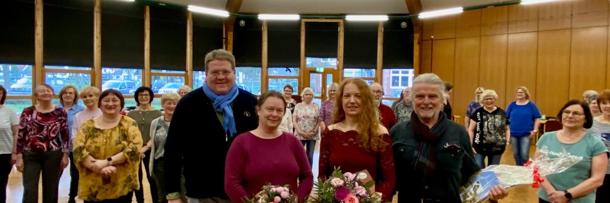 Zahlreiche Erwachsene, die verteilt in einem Veranstaltungsaal stehen und in die Kamera schauen. Im Vordergrund stehen zwei Frauen und zwei Männer, die Blumen oder Präsente in den Händen halten.