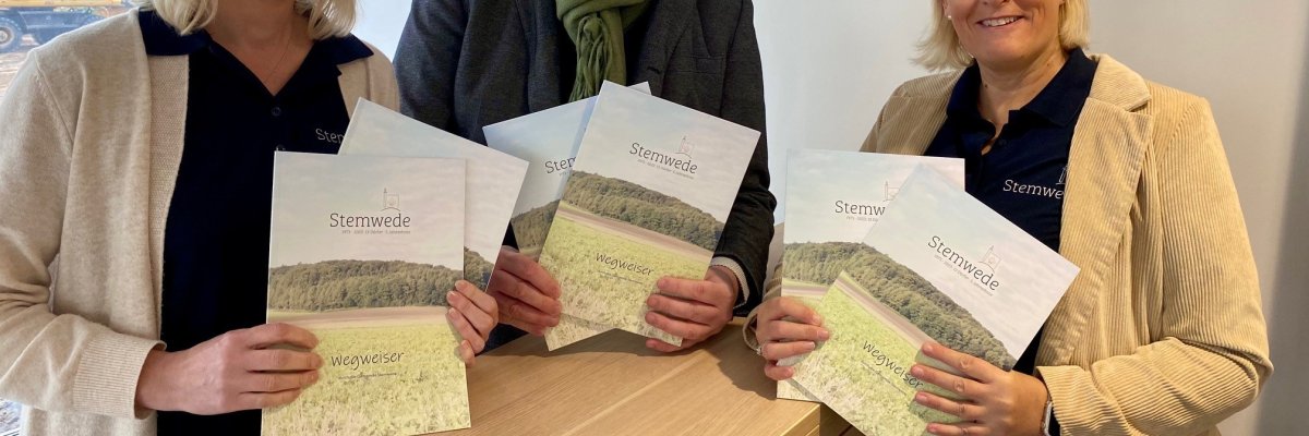 Zwei Frauen, die links und rechts neben einem Mann stehen, halten mehrere Ausgaben einer Broschüre in die Kamera.