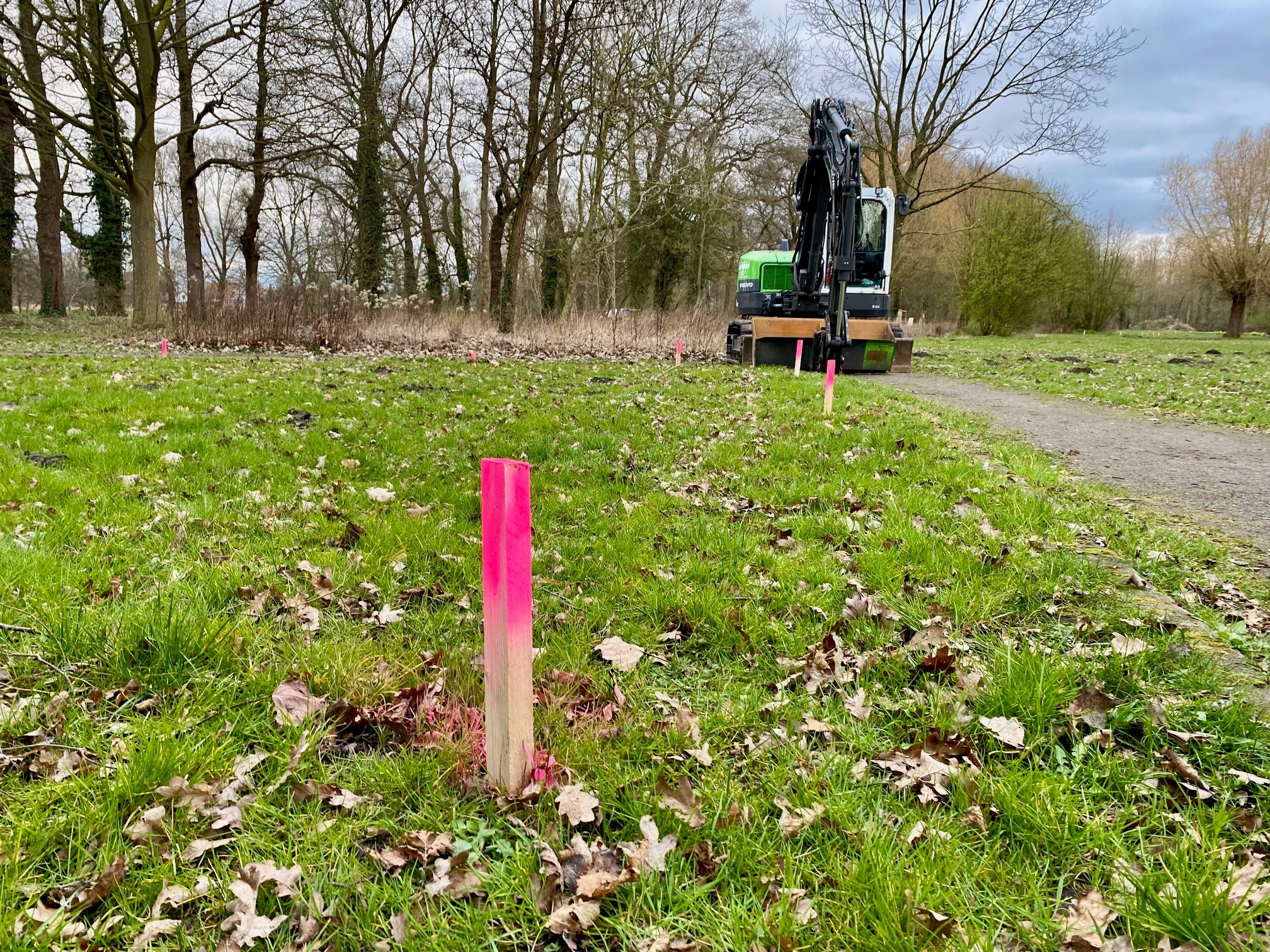 Eine farbige Holzmarkierung steckt in einem Park im Rasen. Im Hintergrund ist ein Bagger zu sehen.