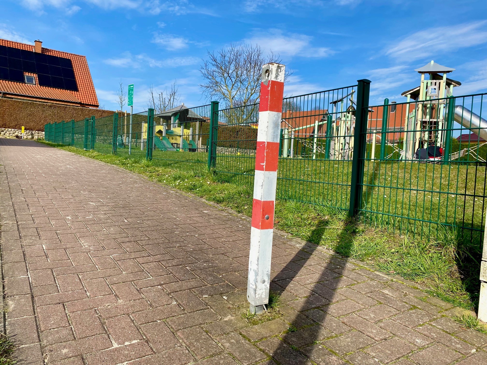Ein rot-weiß gestreifter Absperrpoller auf einem Geh- und Radweg. Im Hntergrund ist ein Spielplatzgelände zu erkennen.