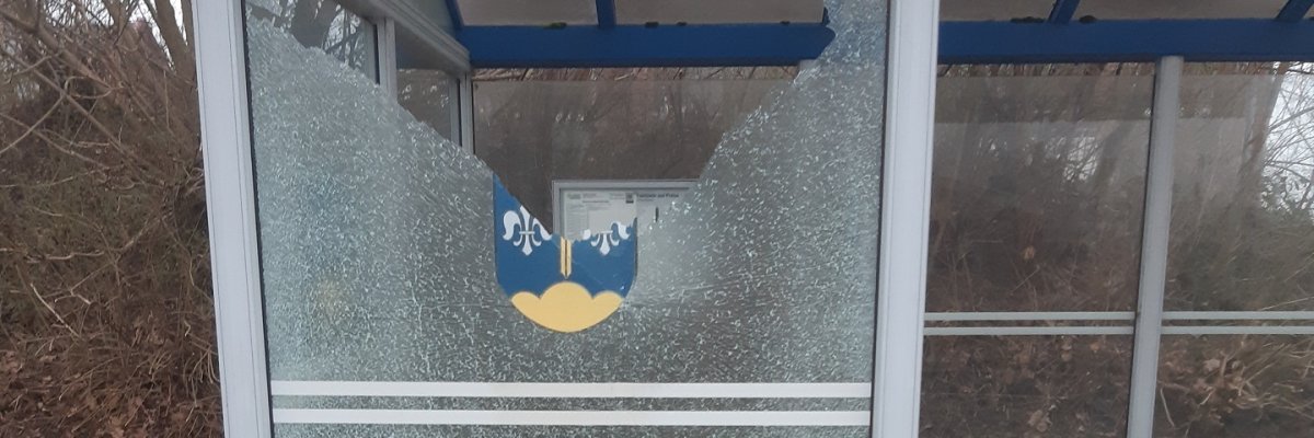 Ein gläsernes Buswartehäuschen, bei dem ein Glaselement zerstört wurde. Die Glasscheibe, auf dem zum Teil noch das Wappen der Gemeinde Stemwede zu sehen ist, ist komplett geborsten und zum Teil zersplittert.