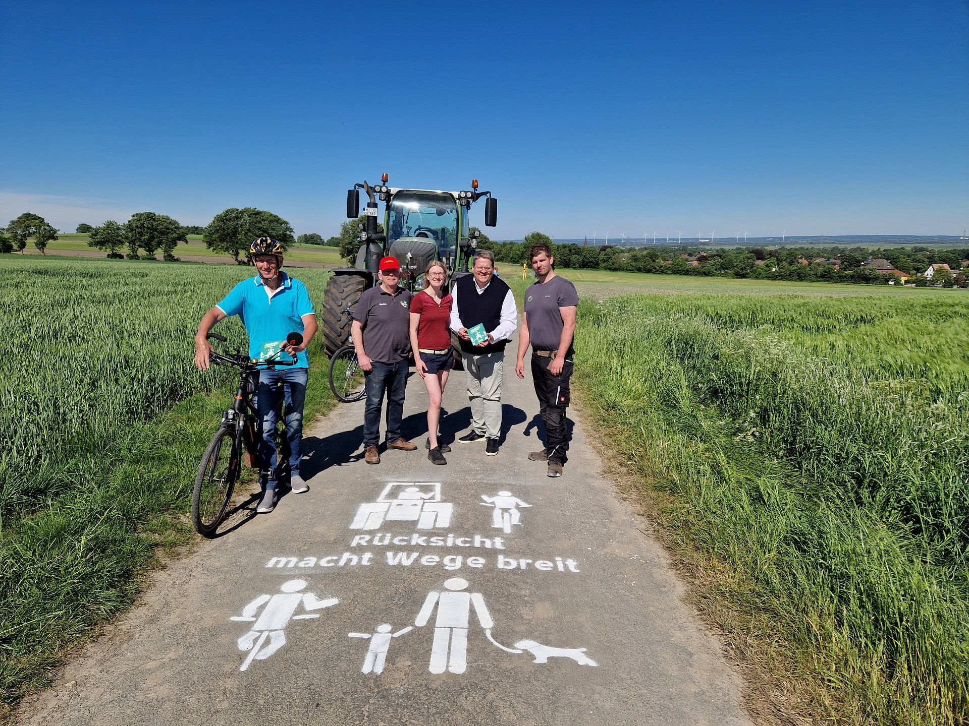 Mehrere Personen stehen bei bestem Wetter auf einem Feldweg zwischen grünen Feldern. Im Hintergrund steht ein grüner Trecker, auf der linken Seite ein Fahrradfahrer und in der MItte der Straße stehen drei Männer und eine junge Frau. Auf die Straße ist mit weißer Farbe der Schriftzug "Rücksicht macht die Wege breit" zu lesen. 