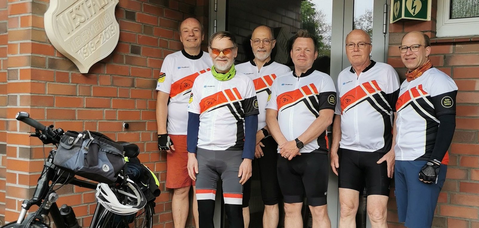 Sechs Männer in Radfahrmontur stehen vor einem Hauseingang. Im linken Teil des Bildes steht ein Fahrrad, an dessen Lenker ein Fahrradhelm hängt.