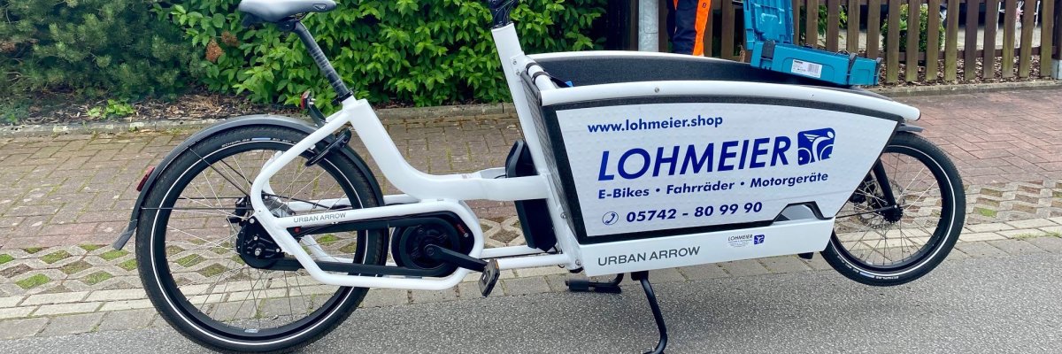 Ein weißes Lasten-E-Bike auf dem der Schriftzug vom Fahrradhaus Lohmeier zu sehen ist, steht an der Straße. Im Hintergrund ist ein Mitarbeiter des Bauhofes zu sehen, der gerade an einer Straßenlaterne arbeitet.