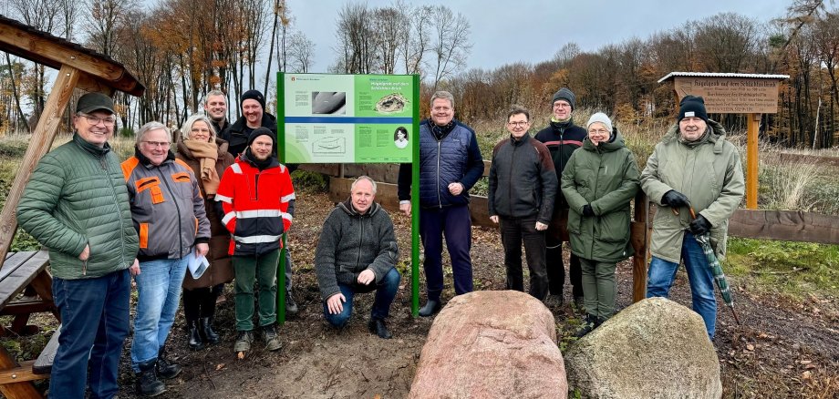 Zehn Männer und zwei Frauen stehen in winterlicher Kleidung im Wald. Zu sehen sind auch zwei Infotafeln, deren Schrift nicht zu entziffern ist, ein Holzunterstand mit Tische und Bank sowie zwei große Findlinge im rechten Vordergrund der Bildes.