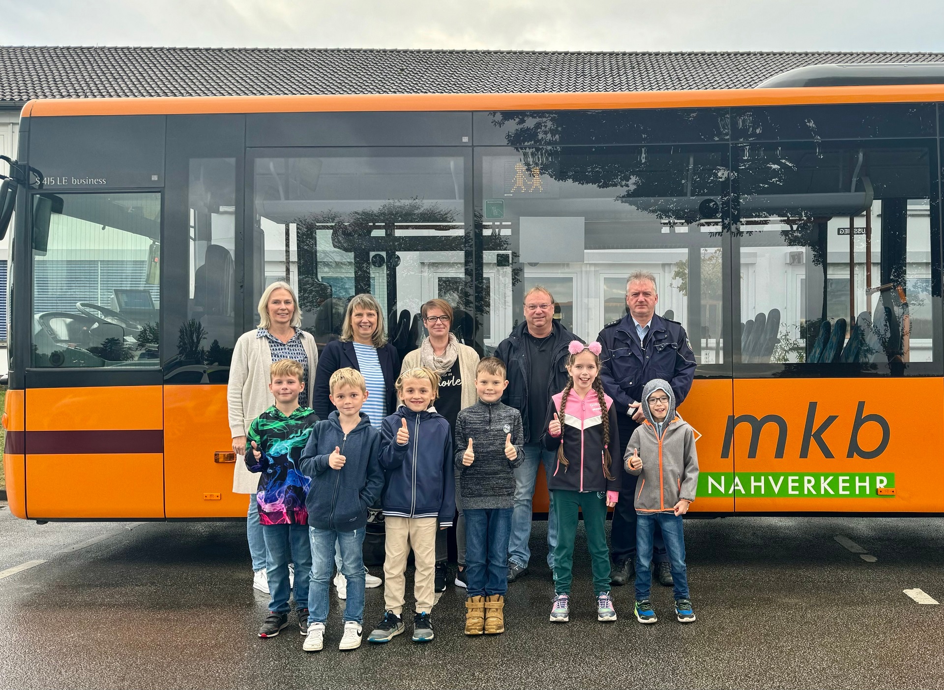 Drei erwachsene Frauen sowie zwei Männer - einer davon ein Polizist - stehen vor einem orangefarbenen Linienbus. Vor den Erwachsenen stehen sechs Kinder - ein Mädchen und fünf Jungs - die den Daumen nach oben zeigen.